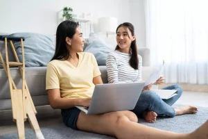Disabled amputee female sitting on ground leaning against couch with support worker looking at laptop